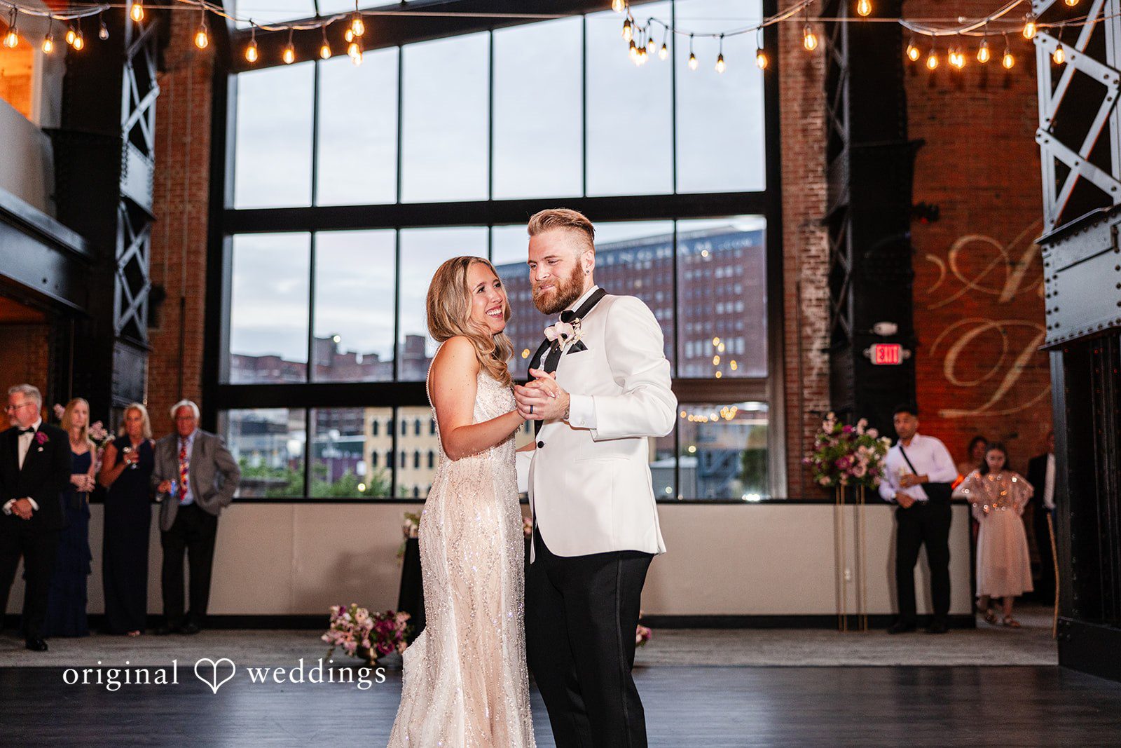 Dan + Alex A bride and groom sharing a dance under hanging lights near a large window view.