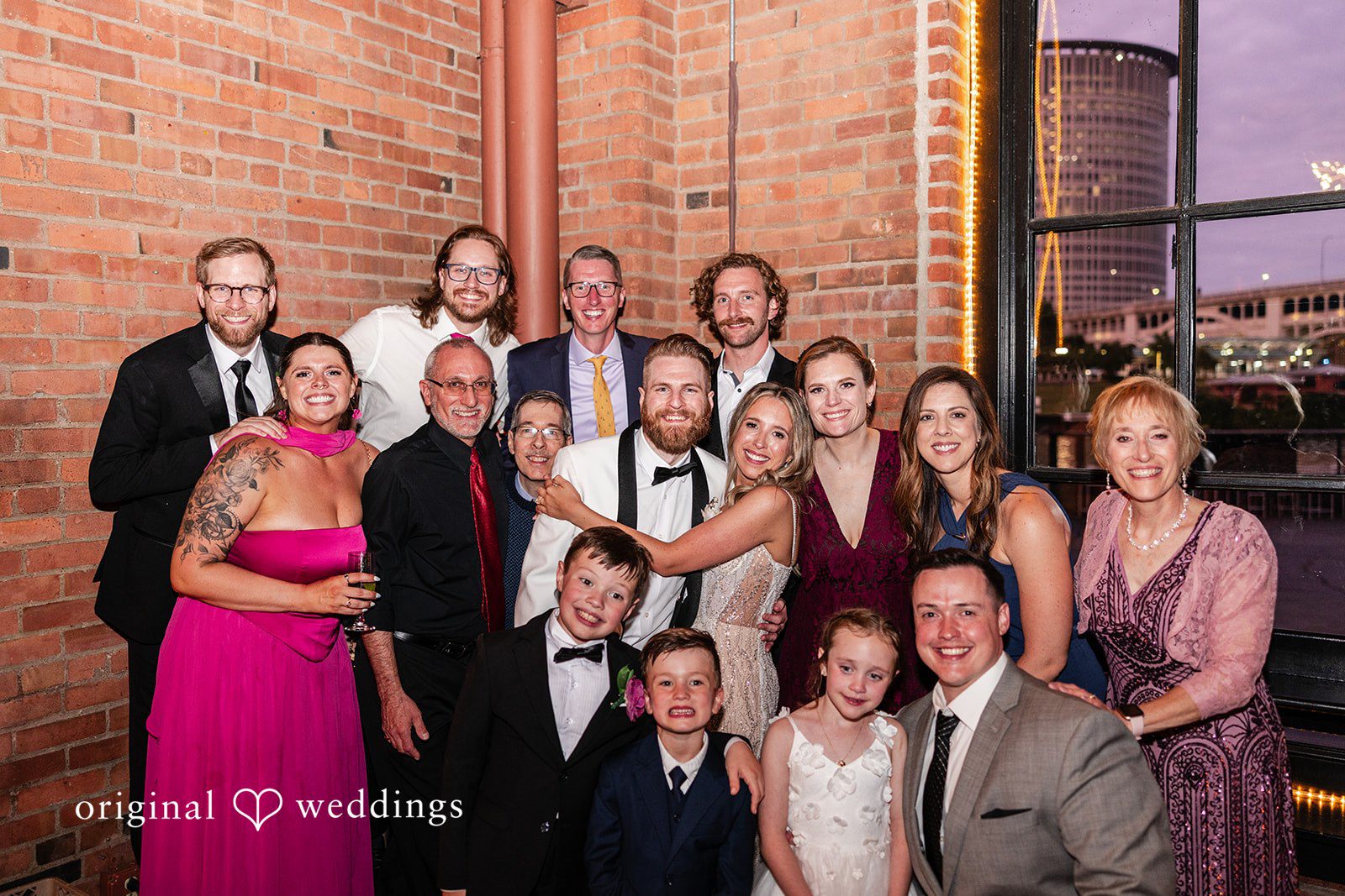 Dan + Alex A large, smiling group of formally dressed wedding guests poses closely around a bride and groom.