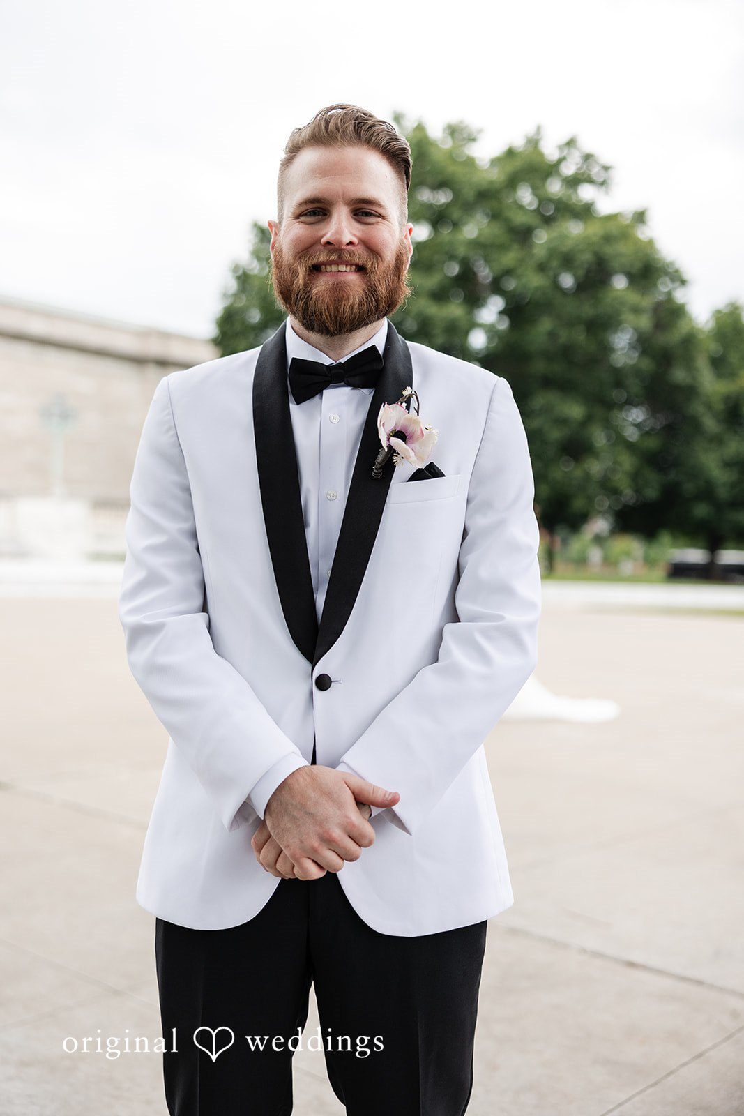 Dan + Alex A smiling groom in a white tuxedo jacket and black bow tie posing against an open green background.