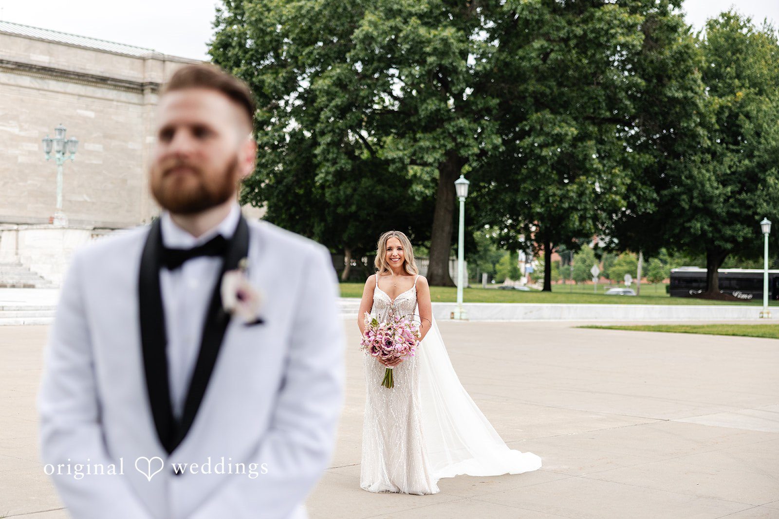 Dan + Alex A groom in a white tuxedo jacket standing in a park while a bride walks in the background.