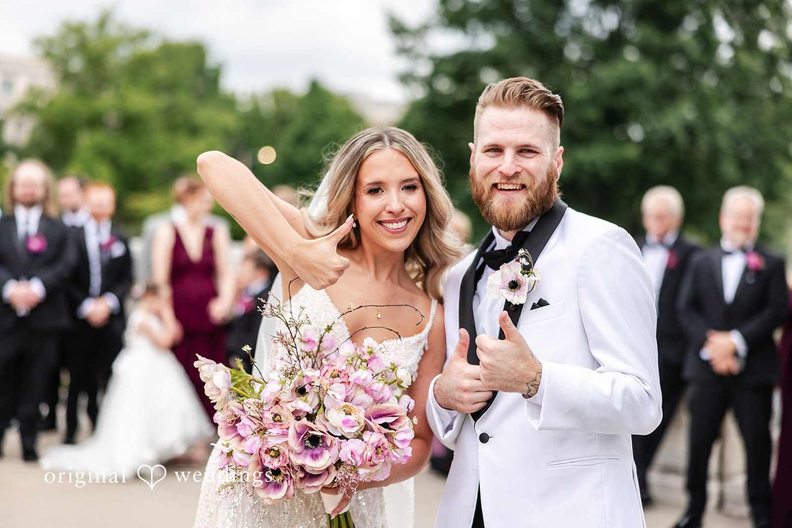 Dan + Alex A joyful bride holding her bouquet high next to a smiling groom in front of greenery.