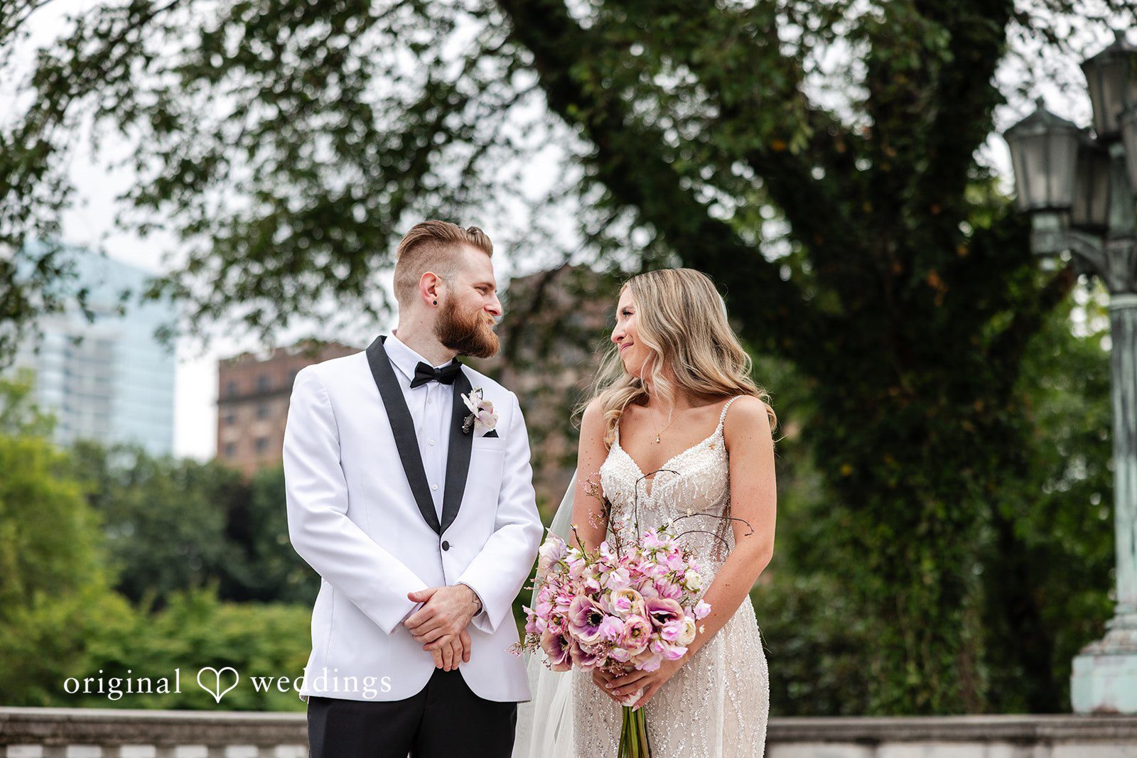 Dan + Alex A groom in a white tuxedo and a bride in a lace gown standing together on a grassy path.