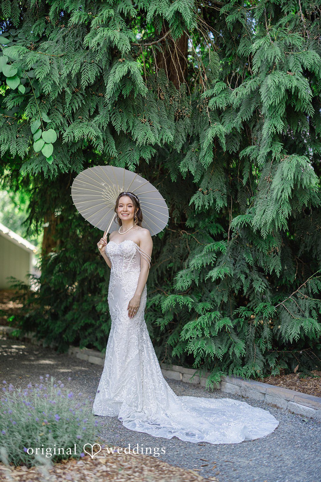 Veronica + Andrew A bride in a long white gown posing with a white parasol on a pathway.