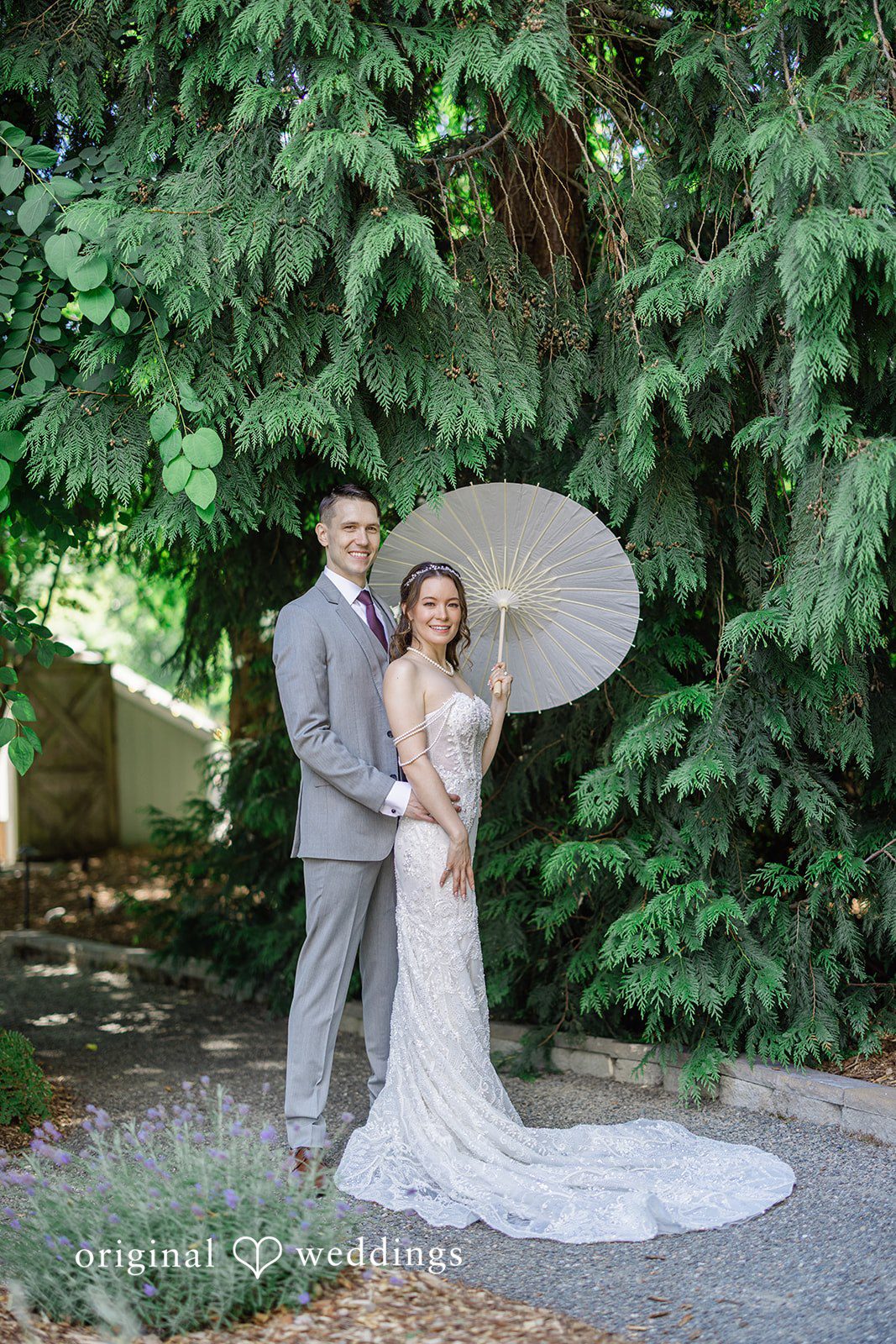 Veronica + Andrew A wedding couple embraces warmly on a path surrounded by green trees.