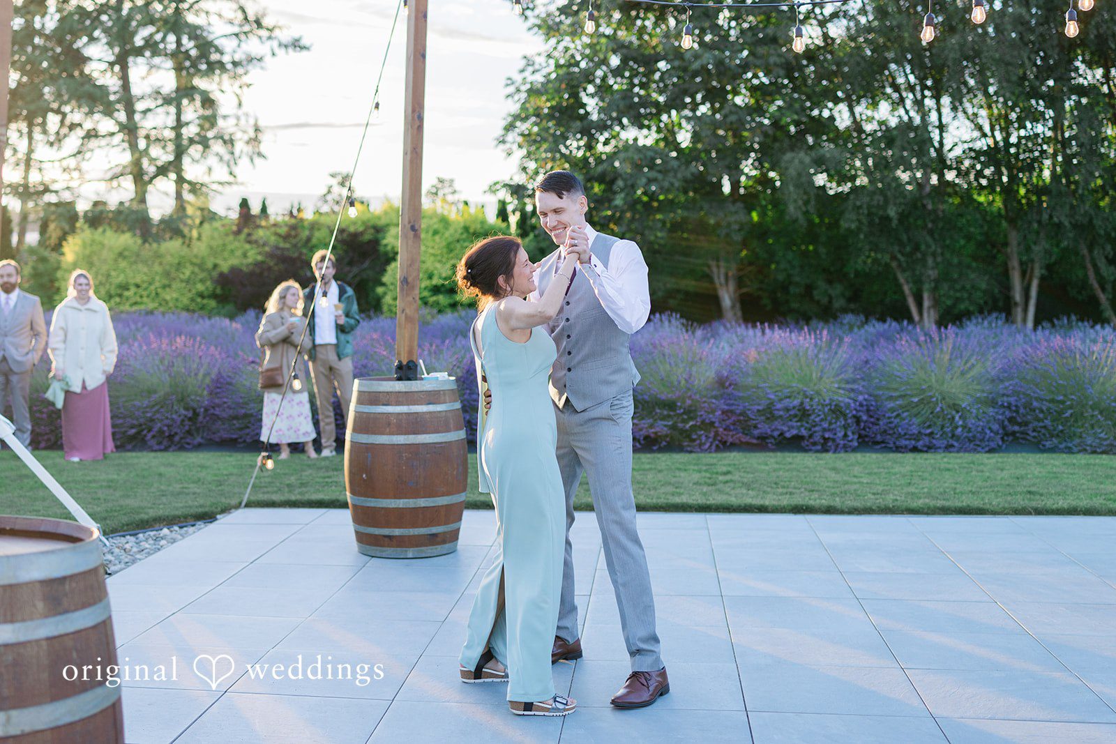Veronica + Andrew A groom in a grey vest and trousers dances on a paved floor with a woman in a light blue jumpsuit.