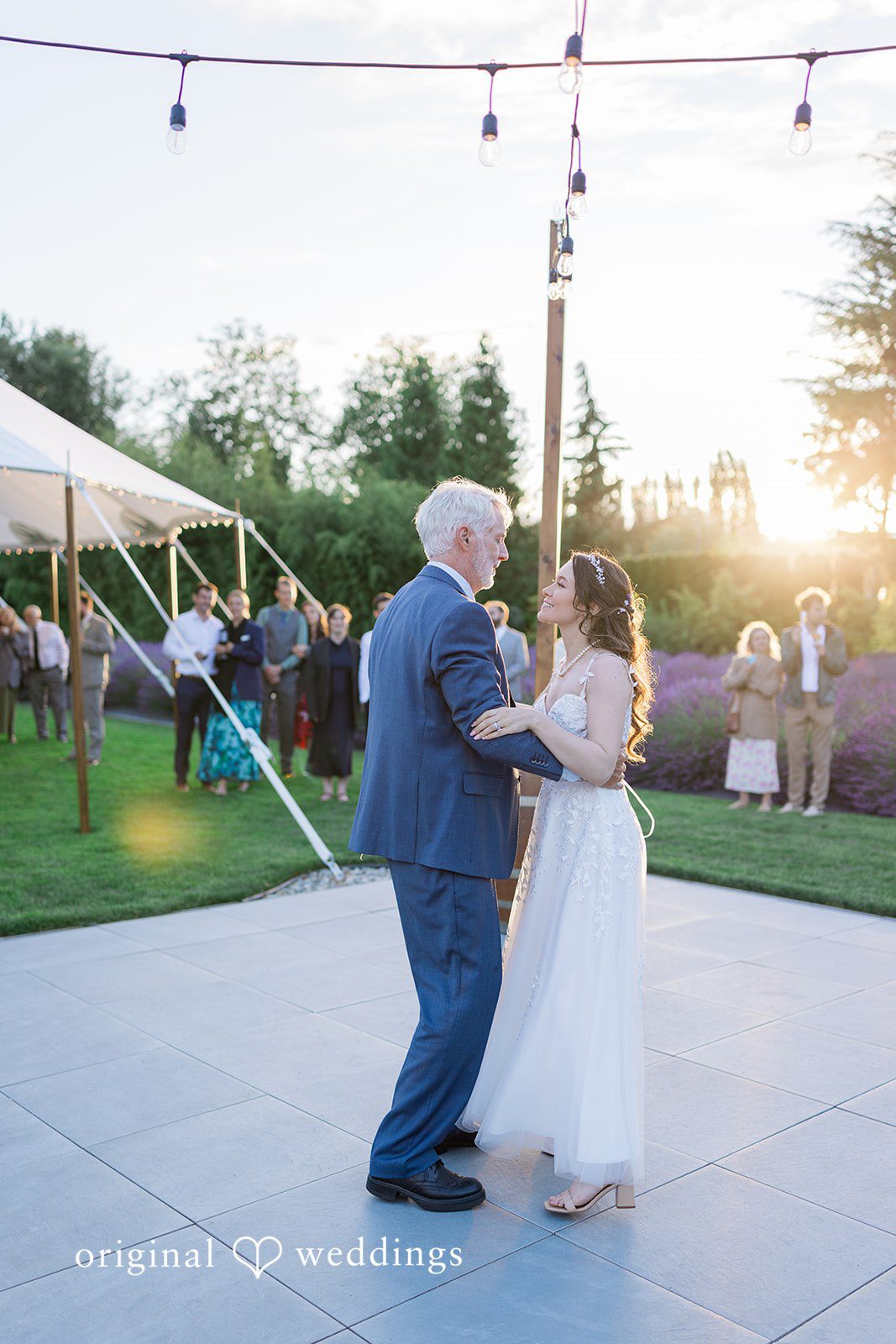 Veronica + Andrew A bride in a white gown dances on an open floor with an older man in a blue suit at sunset.