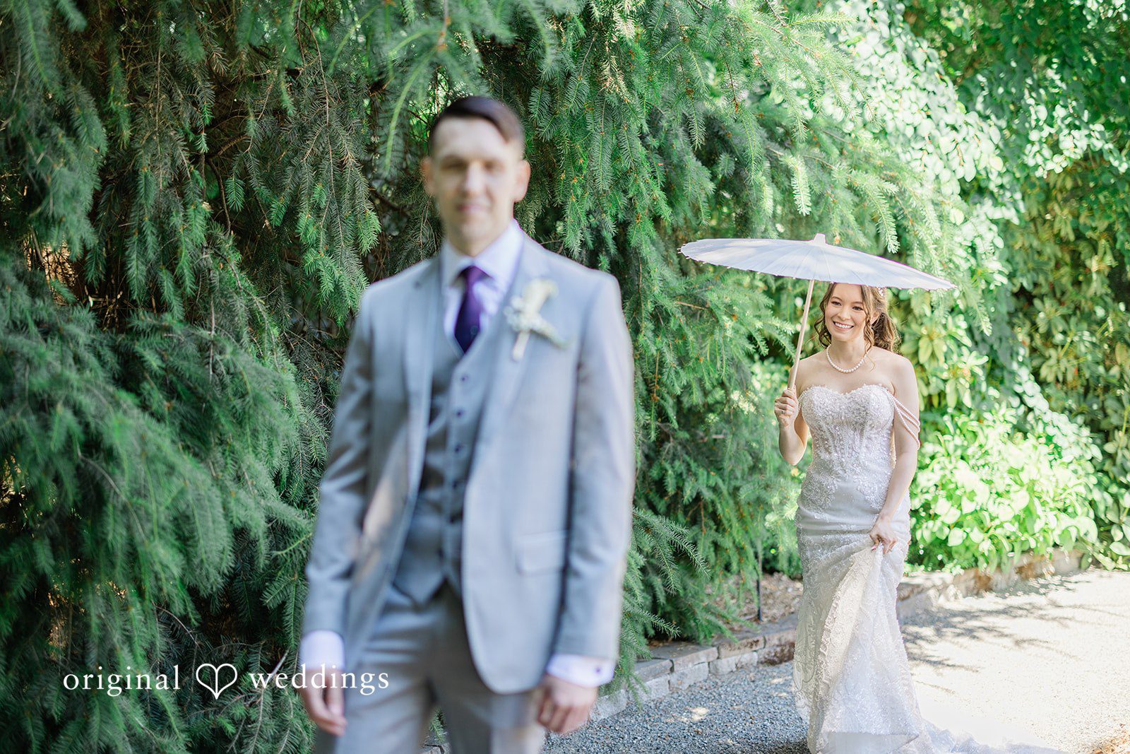 Veronica + Andrew A groom in a grey suit walks ahead of a bride carrying a white parasol.