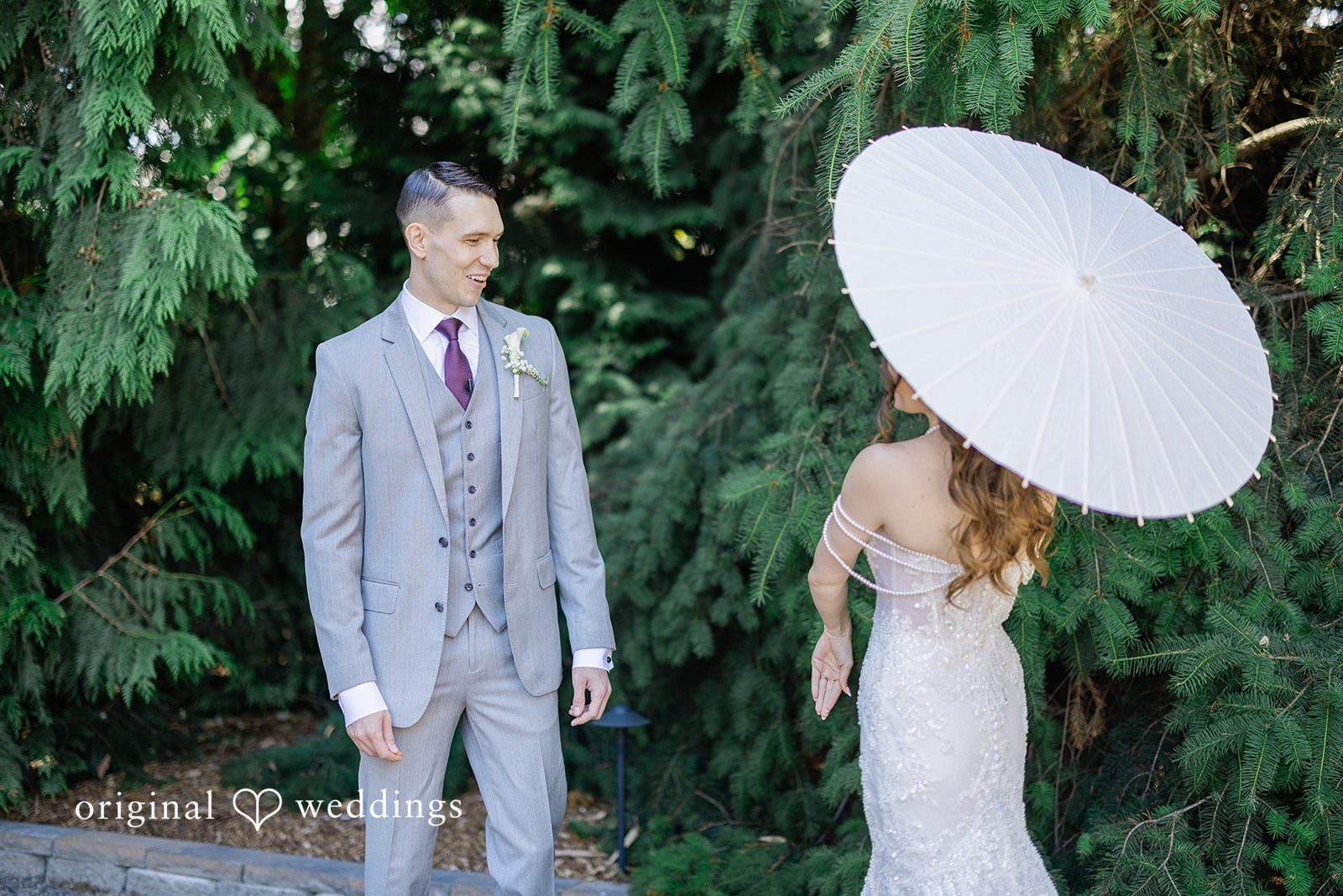 Veronica + Andrew A bride holding a white parasol walks along a path behind the groom.