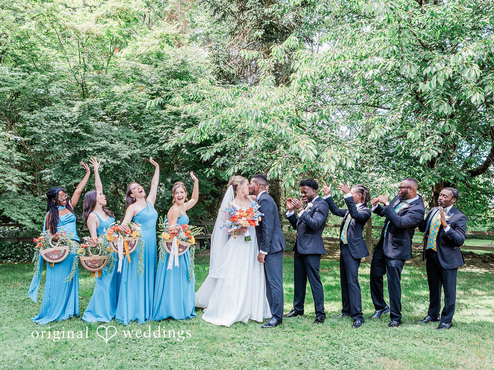 Katrina + Japhet Wedding party standing in a row on a lawn, raising their bouquets and cheering.