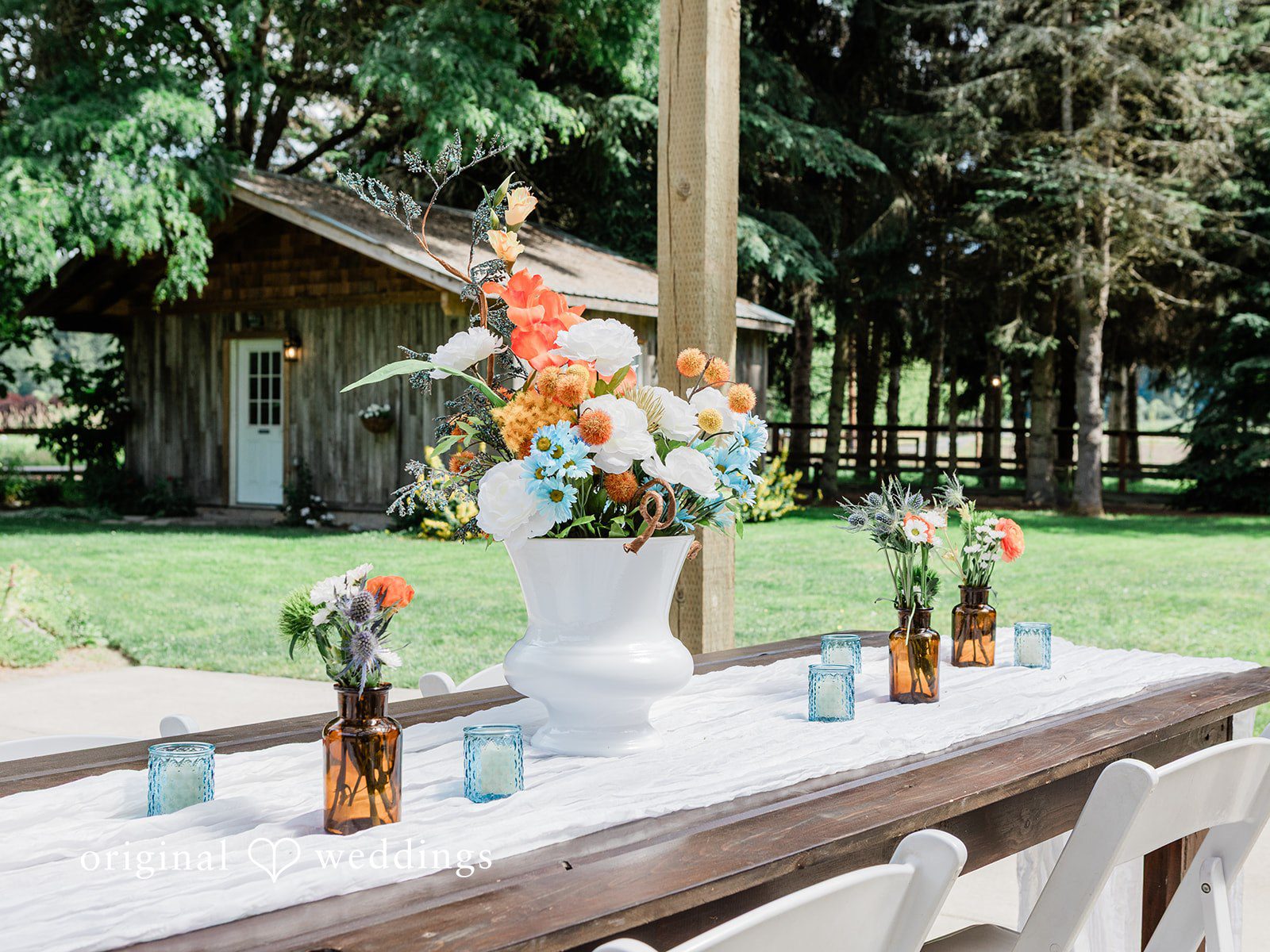 Katrina + Japhet Empty wooden reception table set with small bottles of colorful flowers on a grassy lawn.