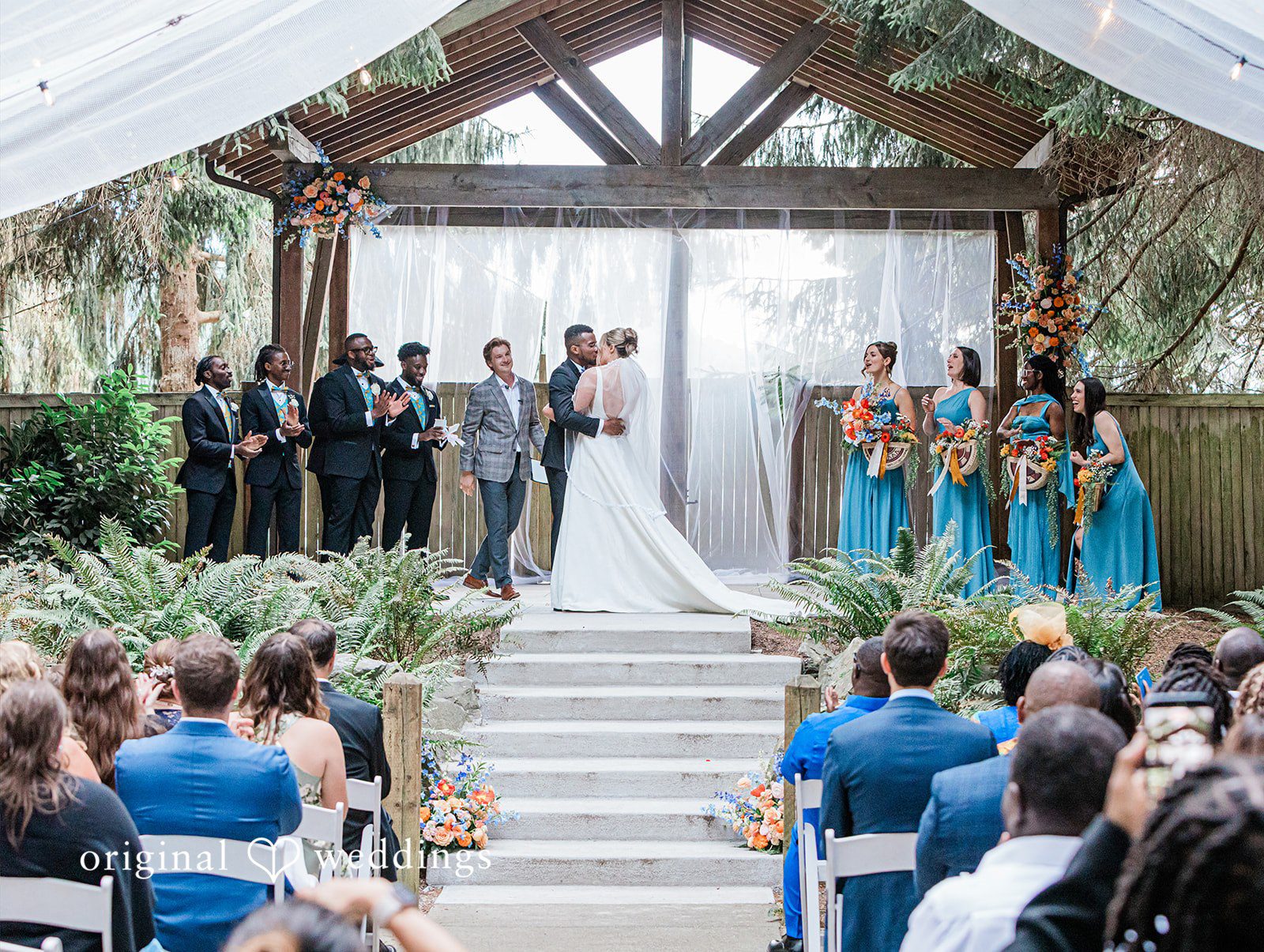Katrina + Japhet Wide view of a wedding ceremony under a timber pavilion filled with guests and wedding party members.