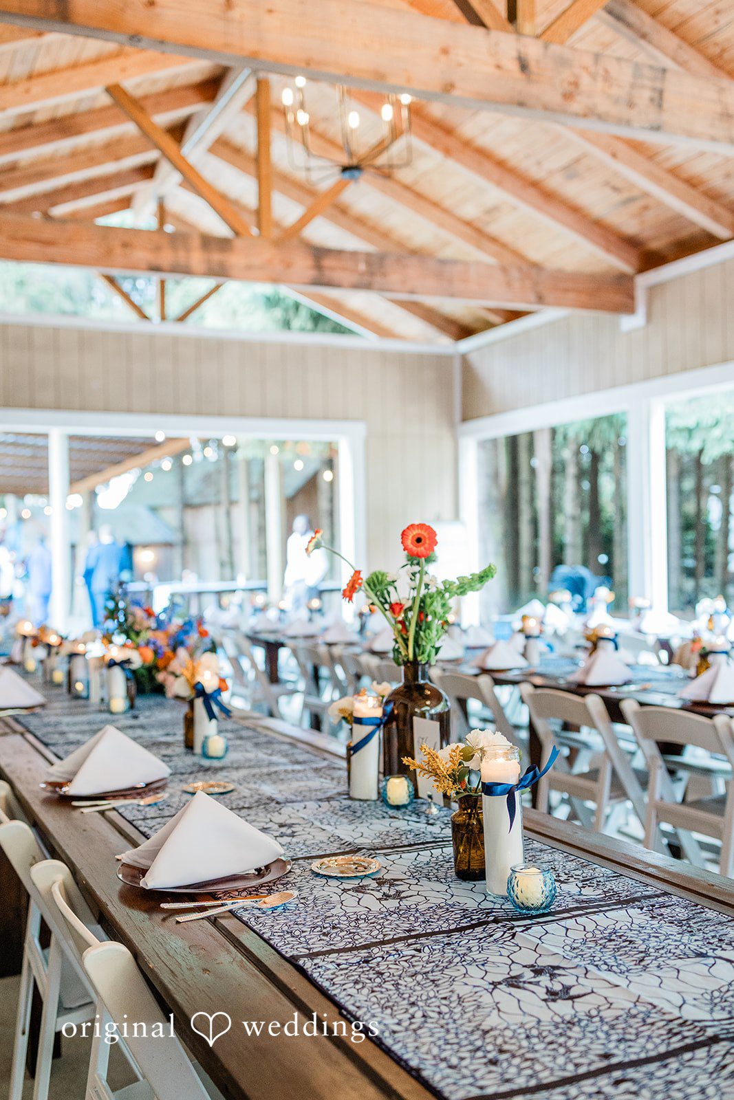 Katrina + Japhet Long wooden dining tables set with blue runners, plates, and floral centerpieces inside a rustic venue.