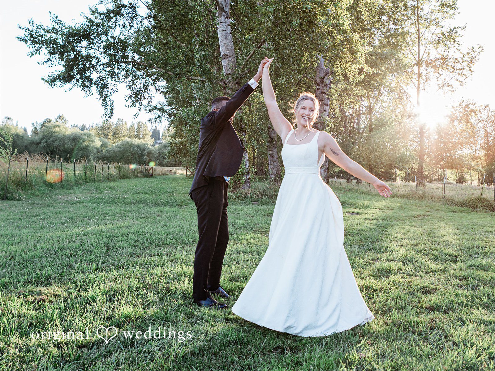Katrina + Japhet Bride and groom holding hands and dancing together in an open grassy field at sunset.