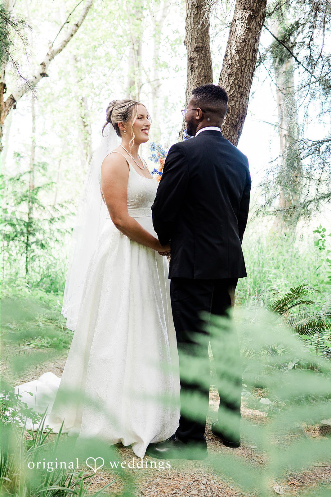 Katrina + Japhet Bride and groom looking at each other while standing on a grassy path surrounded by ferns.