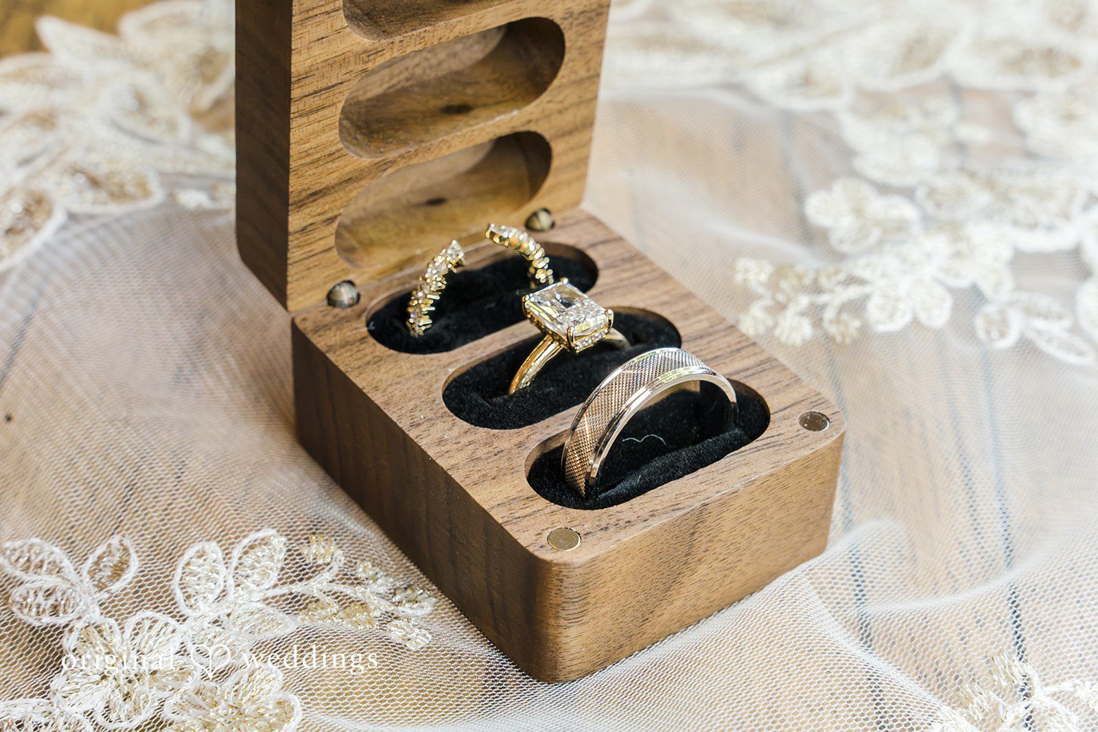 Three elegant rings rest inside individual slots of an open wooden wedding ring box.