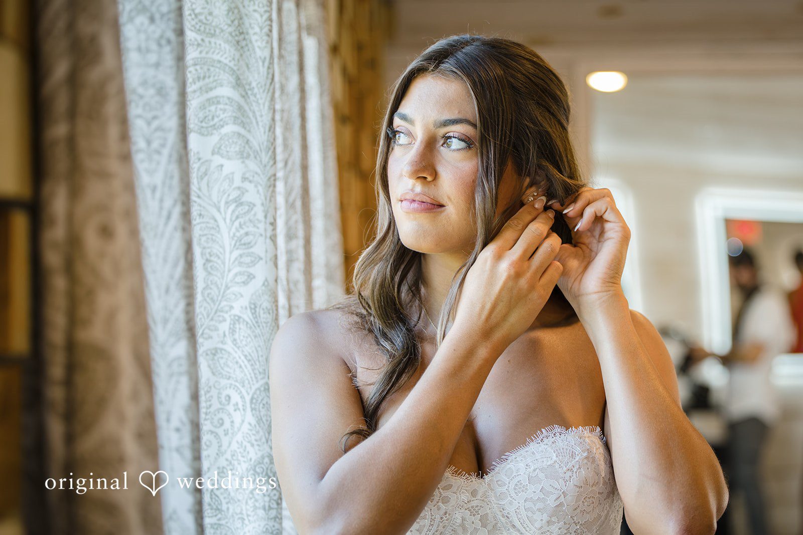 A bride looks to the side while adjusting her earring before a window.