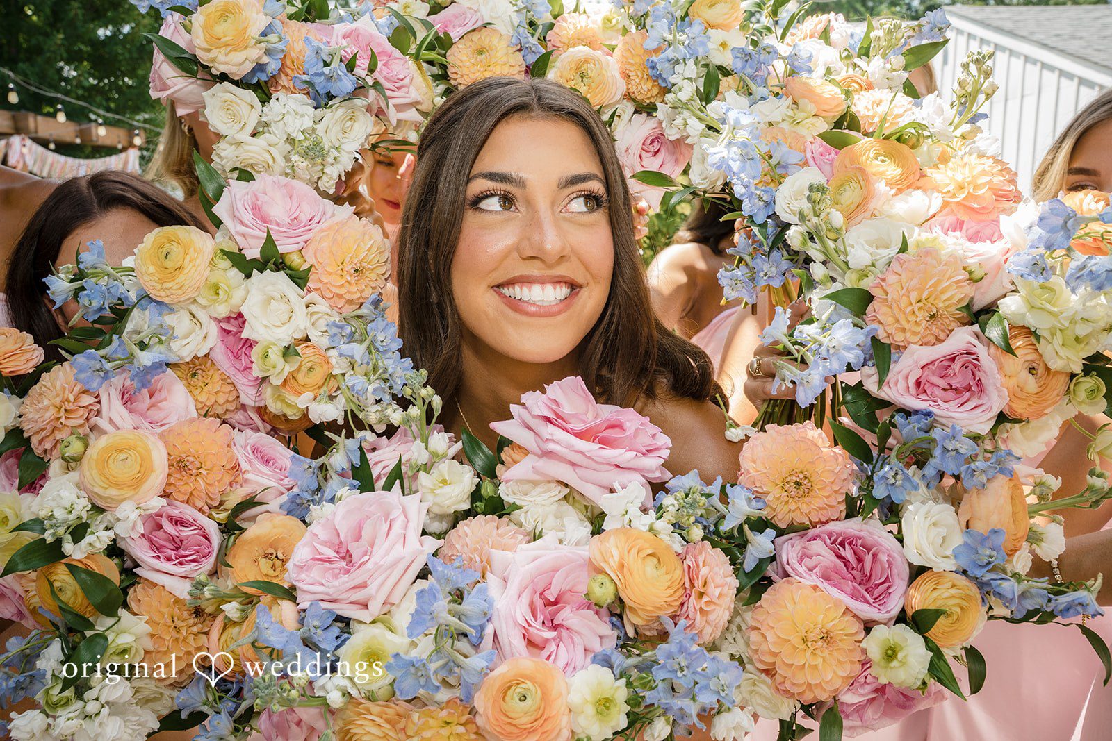 A smiling woman is framed closely behind a vibrant wall of pink, white, and purple flowers.
