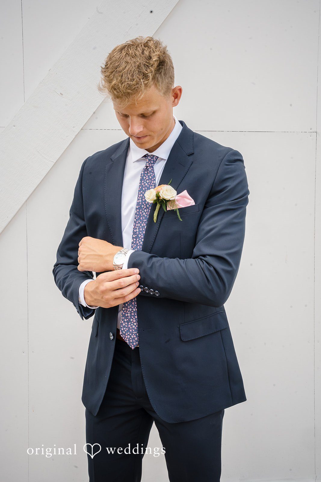 A groom adjusts the sleeve of his dark blue suit jacket against a white backdrop.