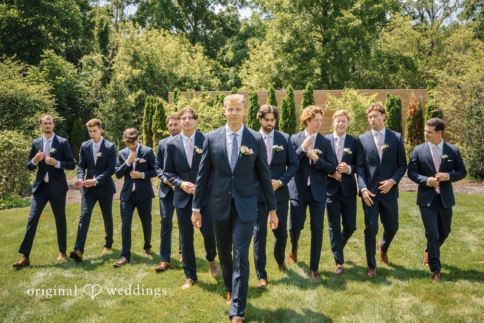 A wedding party in dark formal wear stands lined up across a grassy field.