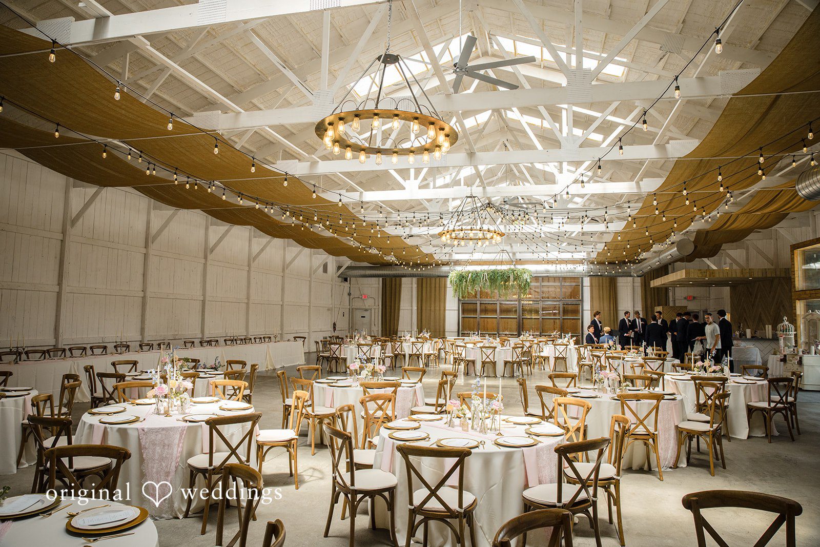 A long rustic reception room features exposed wooden beams, hanging lights, and neatly arranged dining tables.