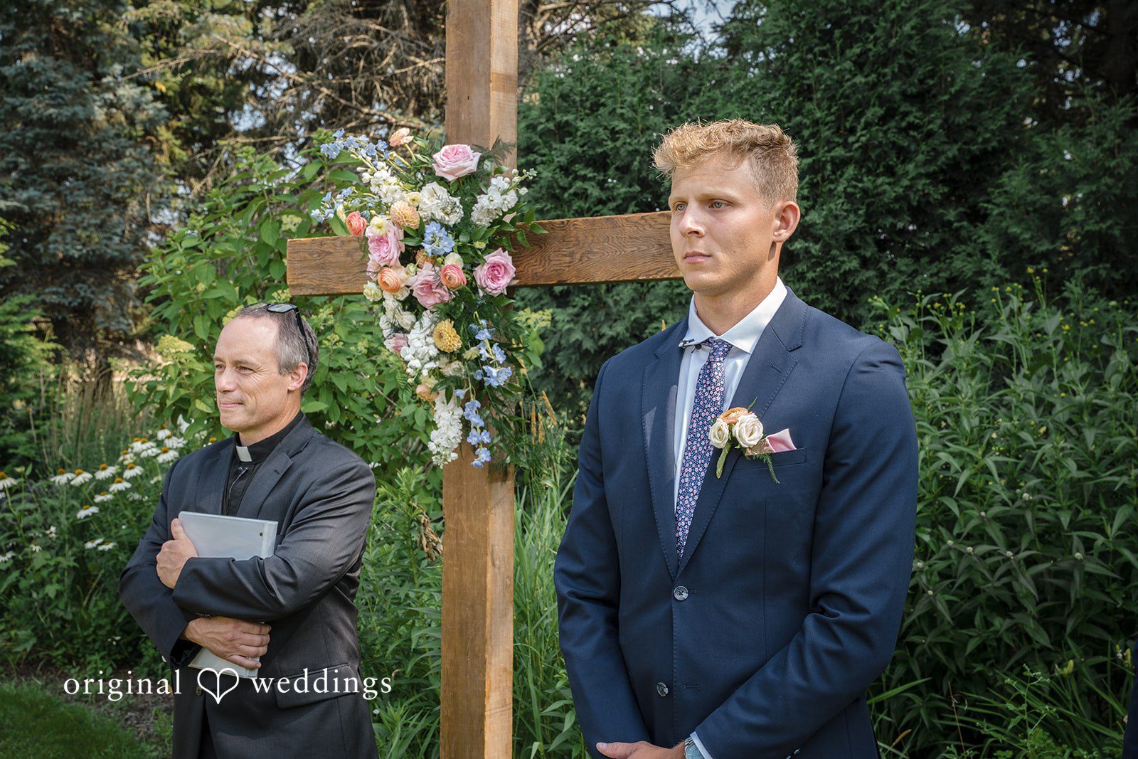 A groom in a blue suit stands next to an officiant before a rustic wooden cross altar.