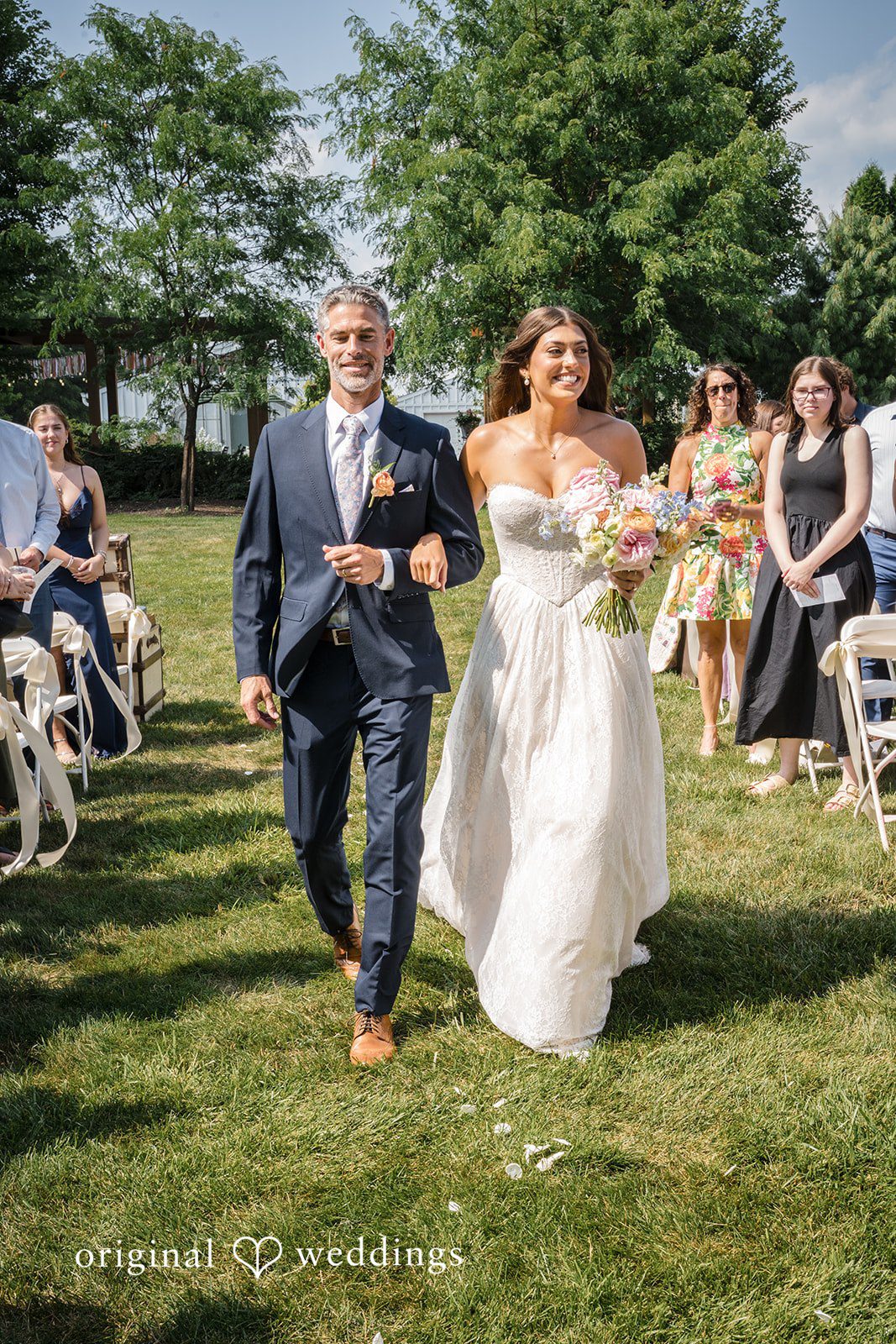 A man walks a bride down a lawn aisle during a ceremony.