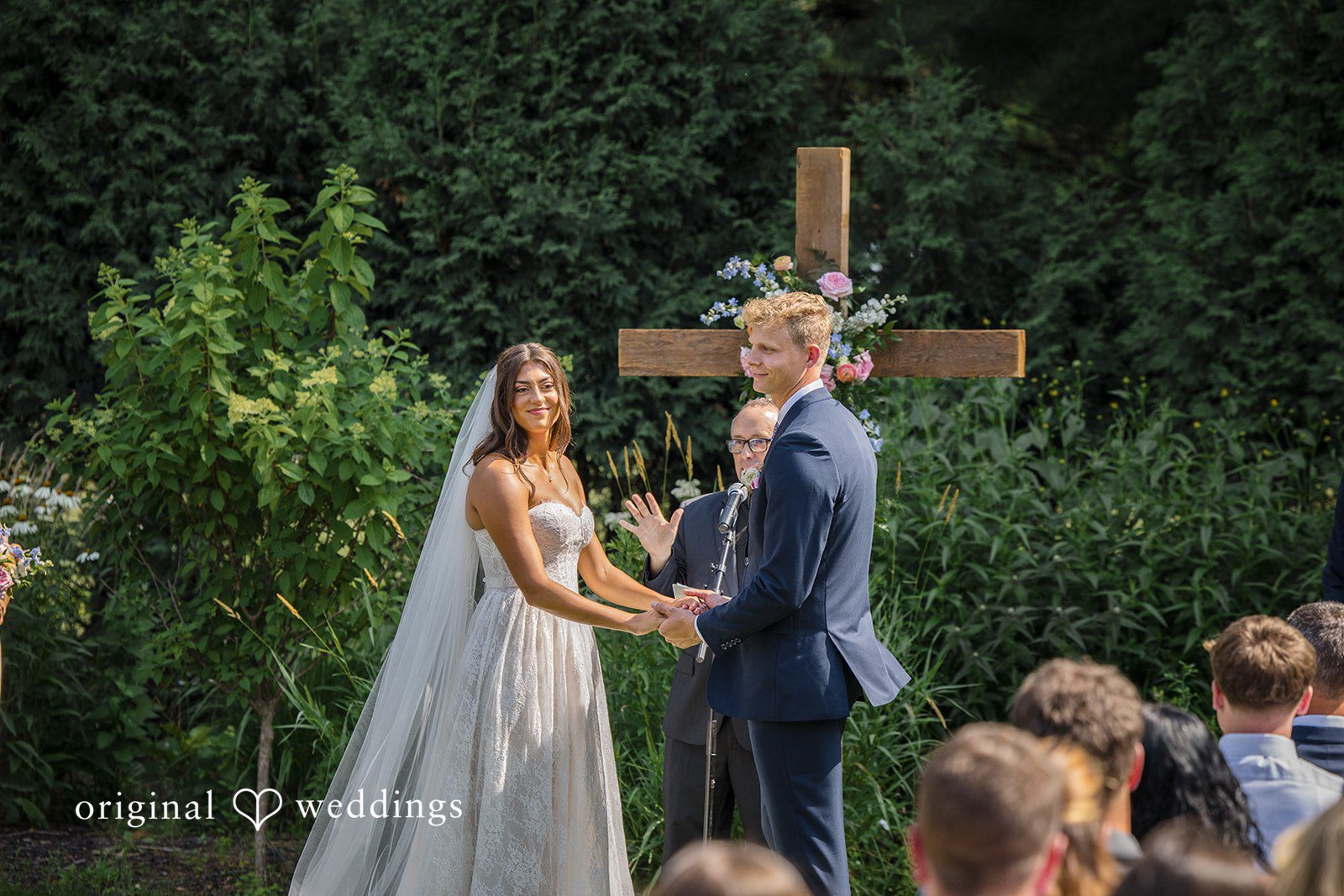 A bride and groom face each other at the altar before a large wooden cross structure.