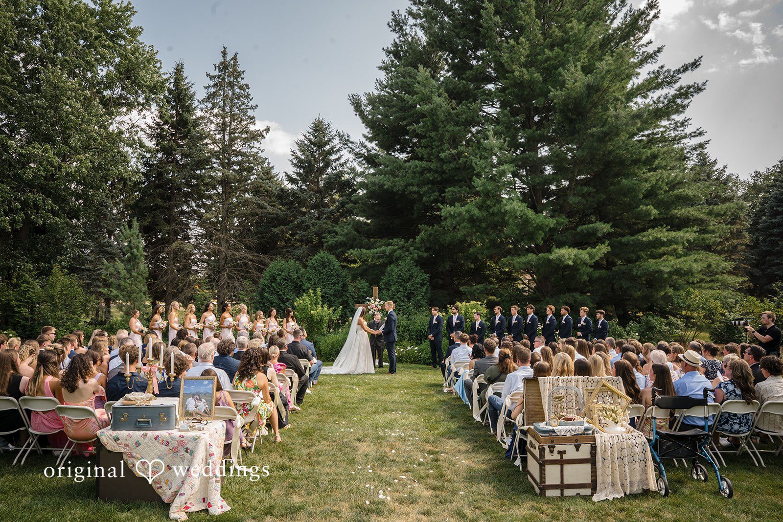 A wide view captures rows of seated wedding guests during an outdoor ceremony in a wooded clearing.