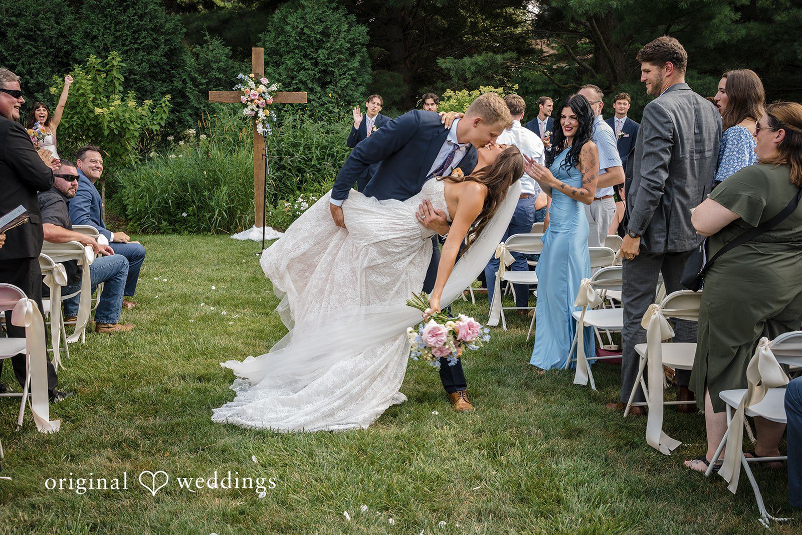A bride walks down a rustic aisle lined with white chairs.