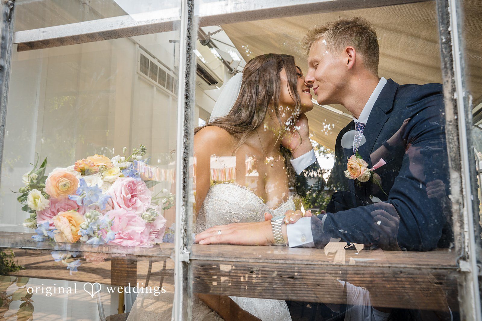 A bride and a groom lean across a small table to share a close, affectionate moment together.