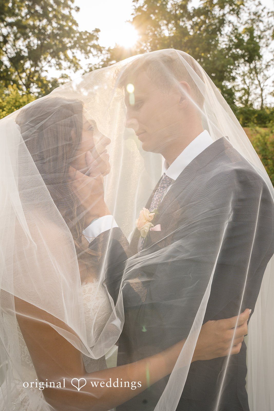 A bride and groom share an intimate embrace while enveloped beneath a flowing sheer veil.