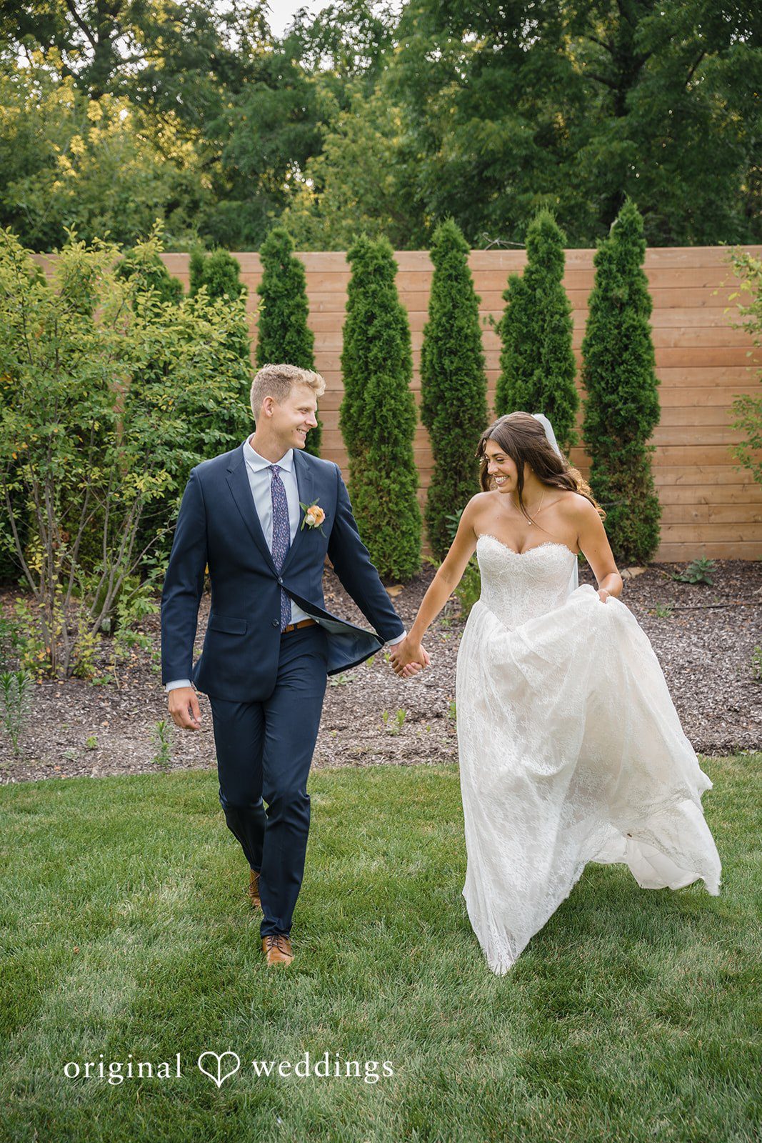 A groom in a dark suit walks hand-in-hand with his bride across a green grassy field.