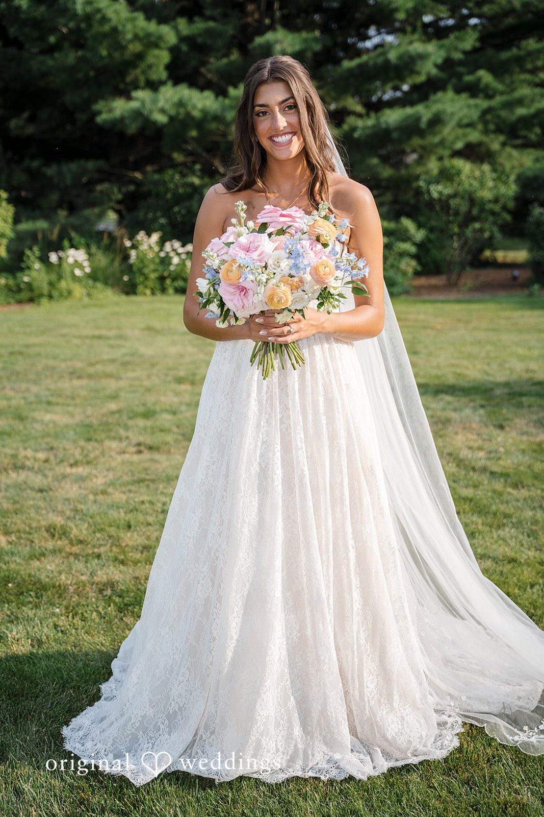 A bride in a flowing white gown poses for a full-length portrait on a grassy lawn.