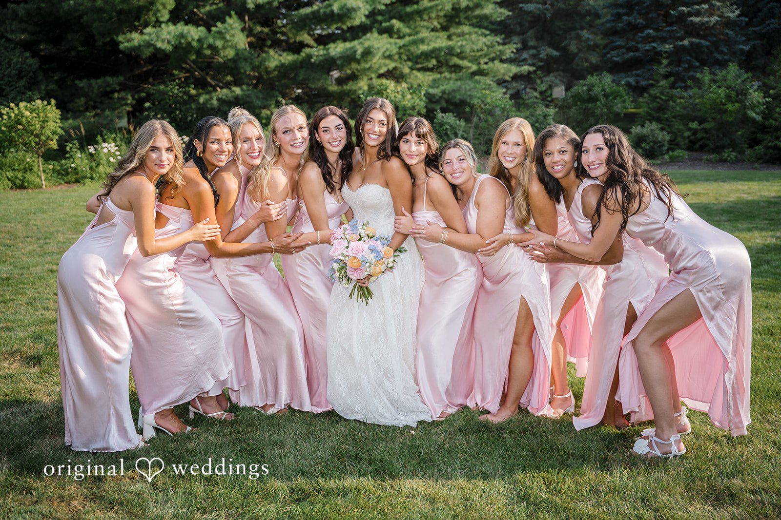 A bridal party dressed in light pink gowns poses together on a grassy field.
