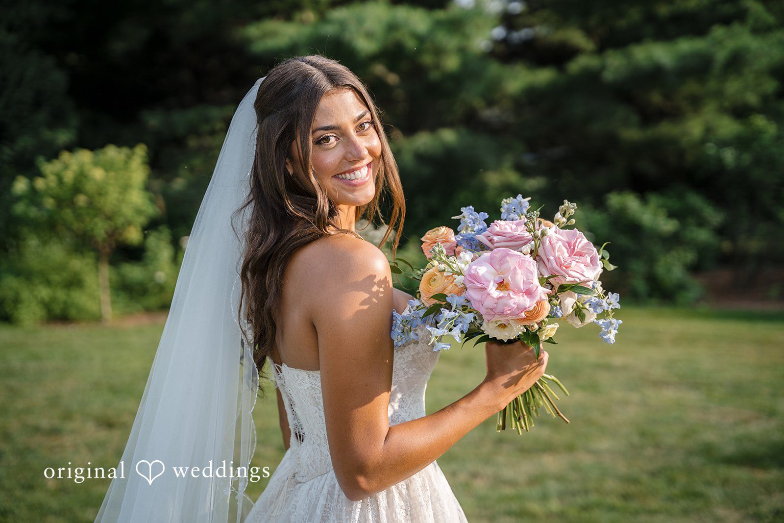 A bride with a long sheer veil smiles while holding her colorful bouquet among green foliage.