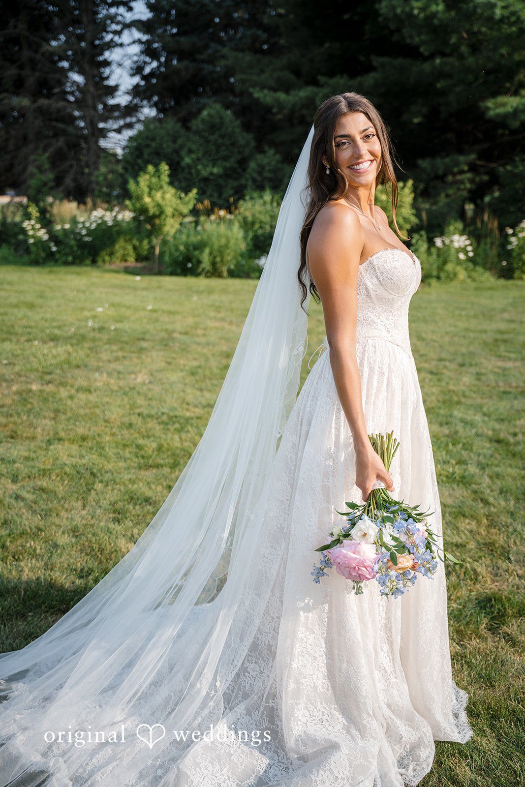 A bride walks across a lawn while her long white train trails behind her.