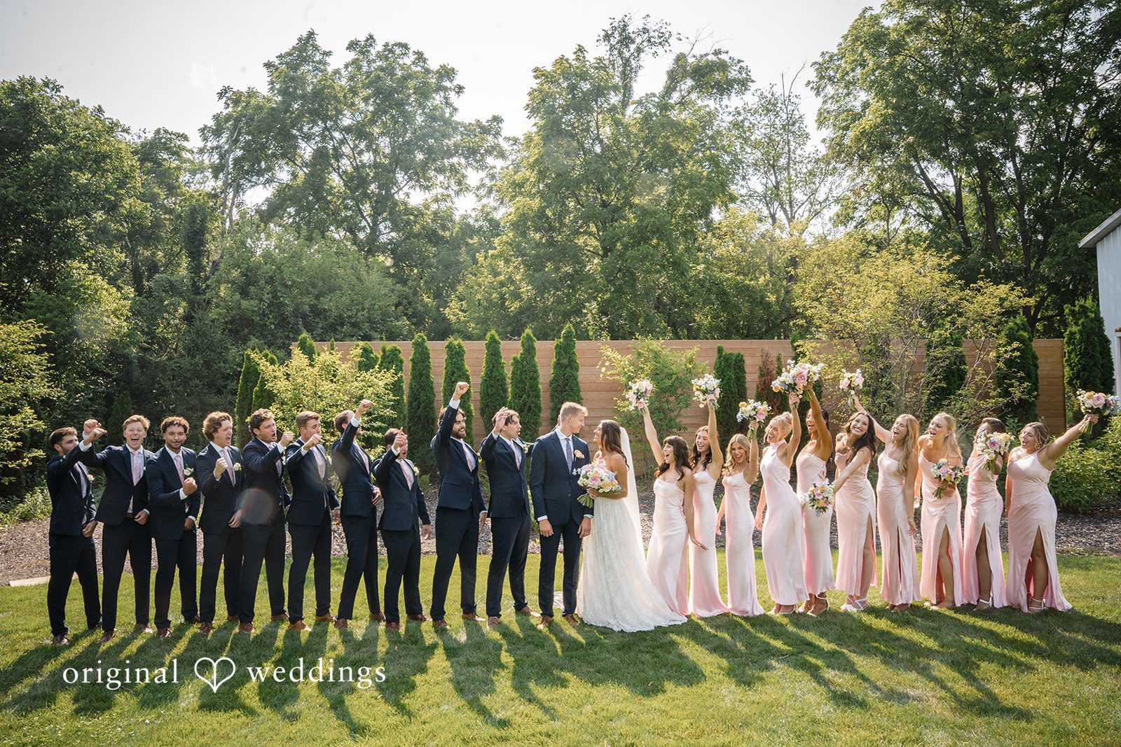 A large group wedding portrait shows attendants and guests gathered on a green lawn with background trees.