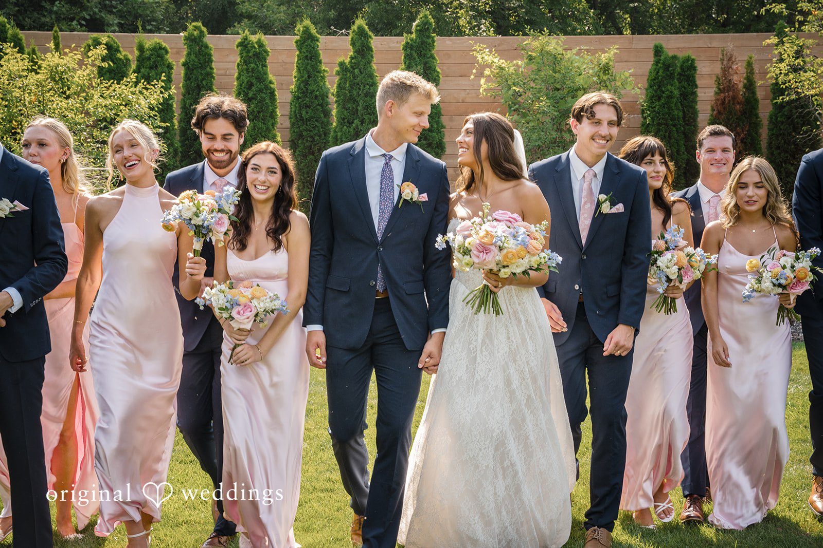 A wedding party stands together in a row along a rustic wooden wall.