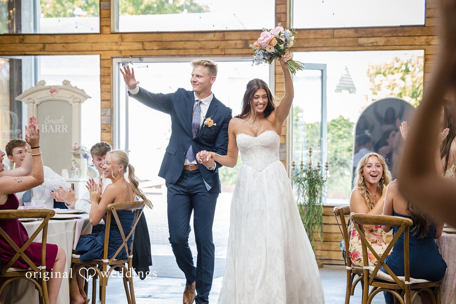 A bride in a white gown walks across a rustic room filled with wedding guests.