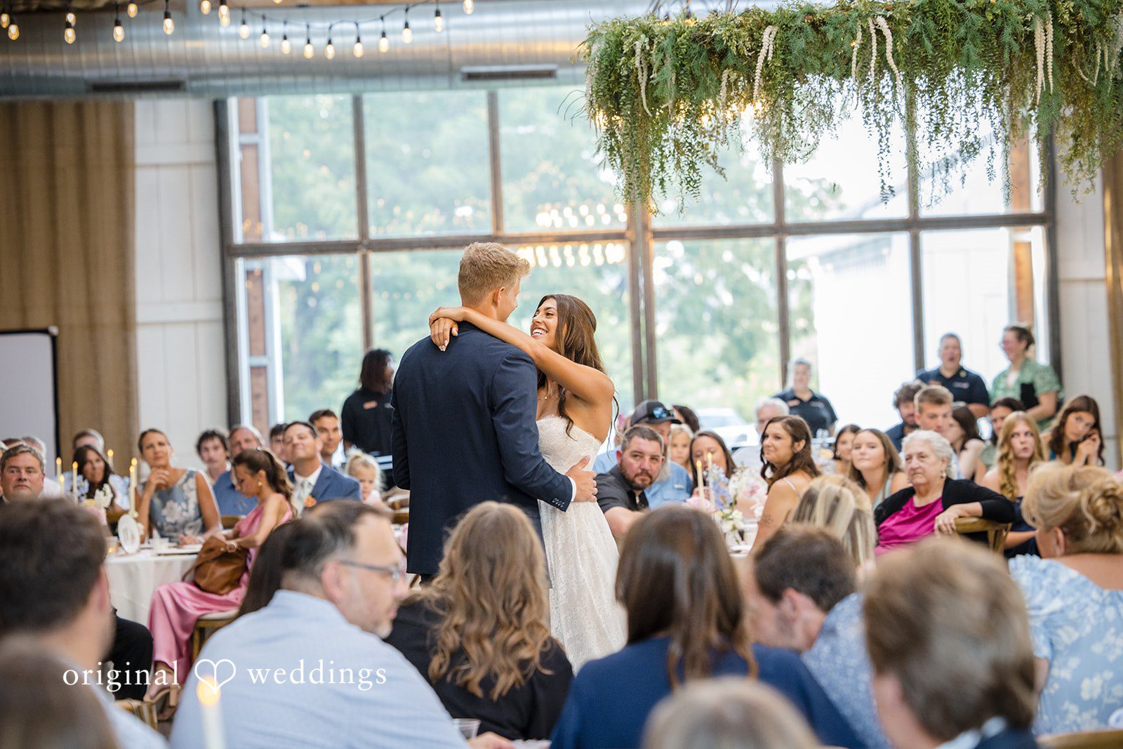 A group of wedding reception guests sit at long tables looking toward a focal point.