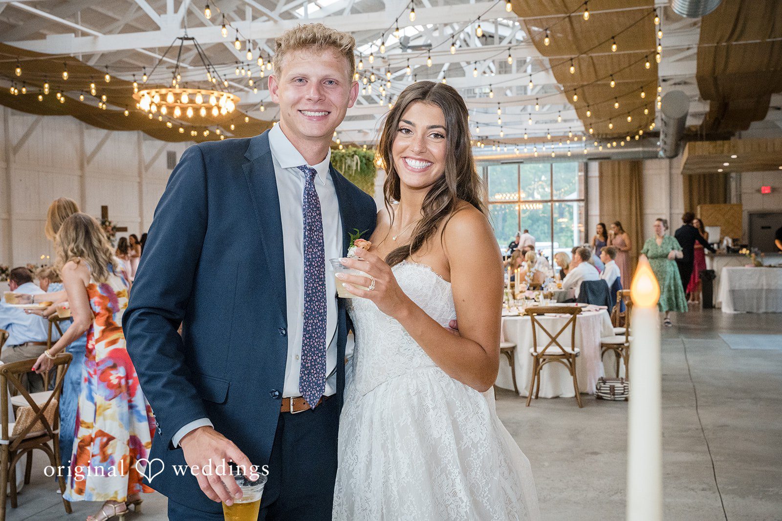 A groom in a dark suit stands beside his smiling bride near a brightly lit entryway.