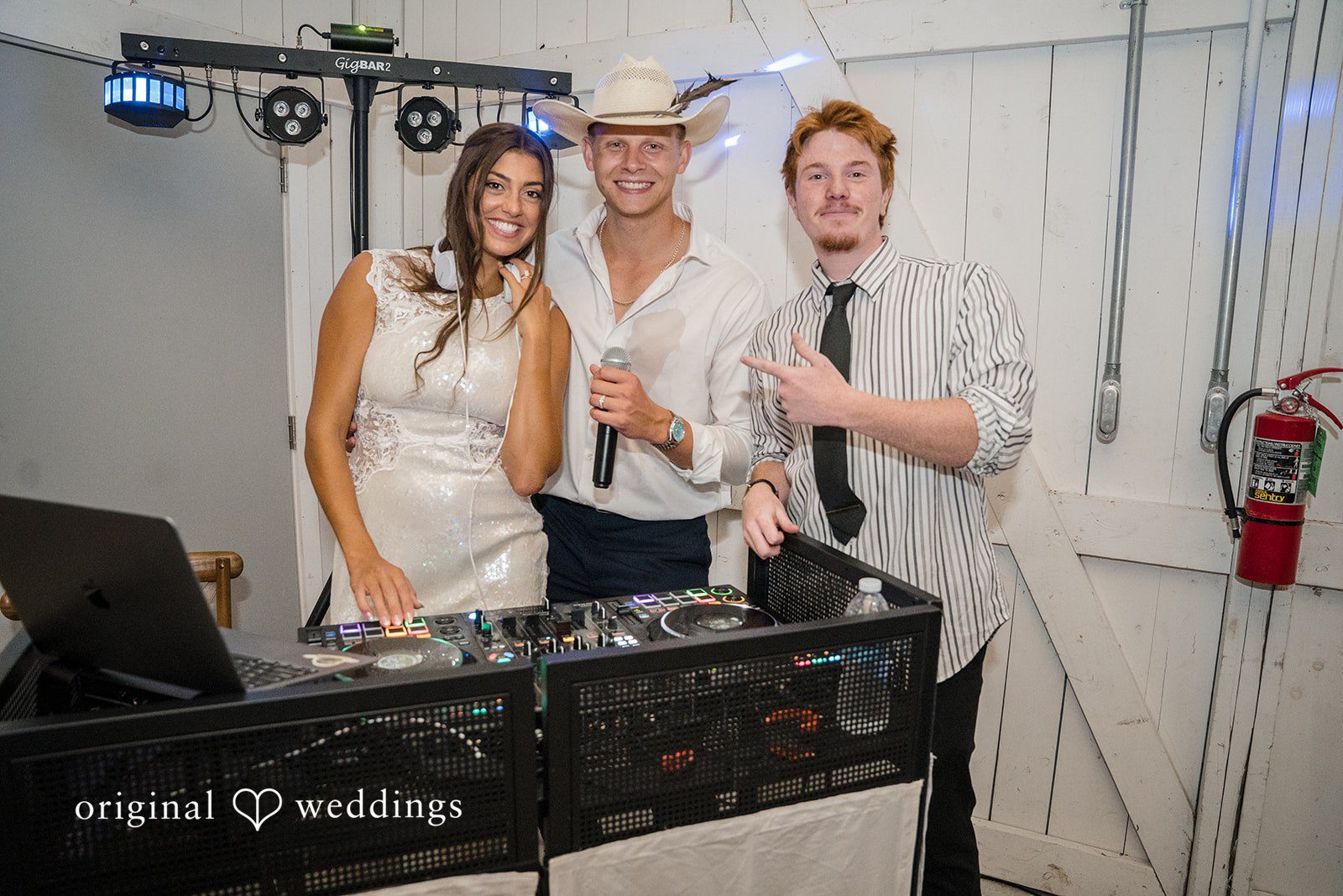 A bride and groom stand behind a DJ booth table equipped with music gear.