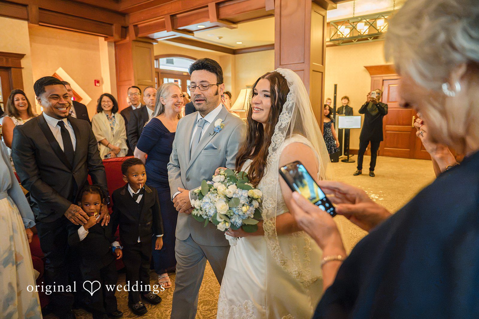 Ava + Zach A bride and groom walking together through a crowded hallway while a guest takes a picture with a smartphone.