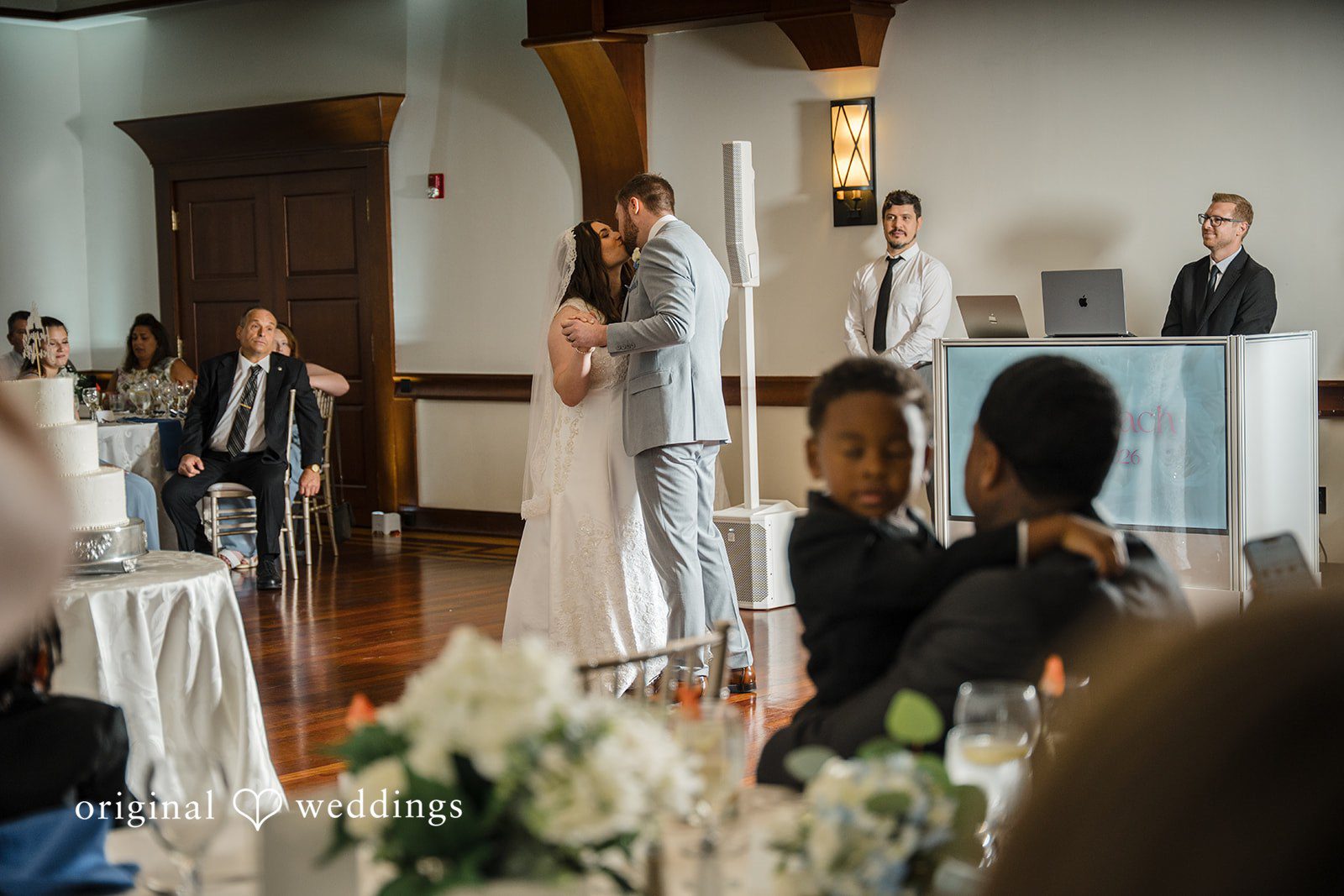 Ava + Zach A bride in a white gown and a groom in a light gray suit sharing a kiss on the dance floor.