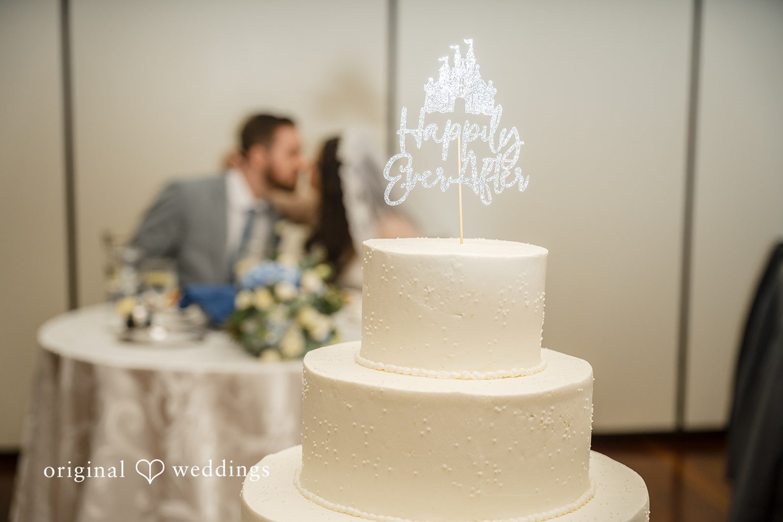 Ava + Zach A three-tiered white wedding cake decorated with flowers sitting on a reception table.