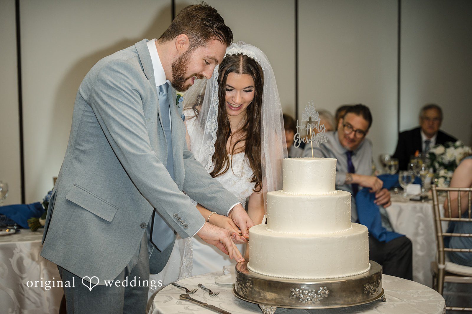 Ava + Zach A bride and groom cutting their three-tiered white wedding cake.