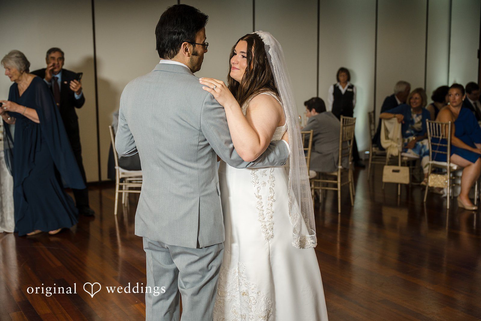Ava + Zach A bride in an embellished white gown and long veil sharing a slow dance with a groom in a gray suit while guests look on.