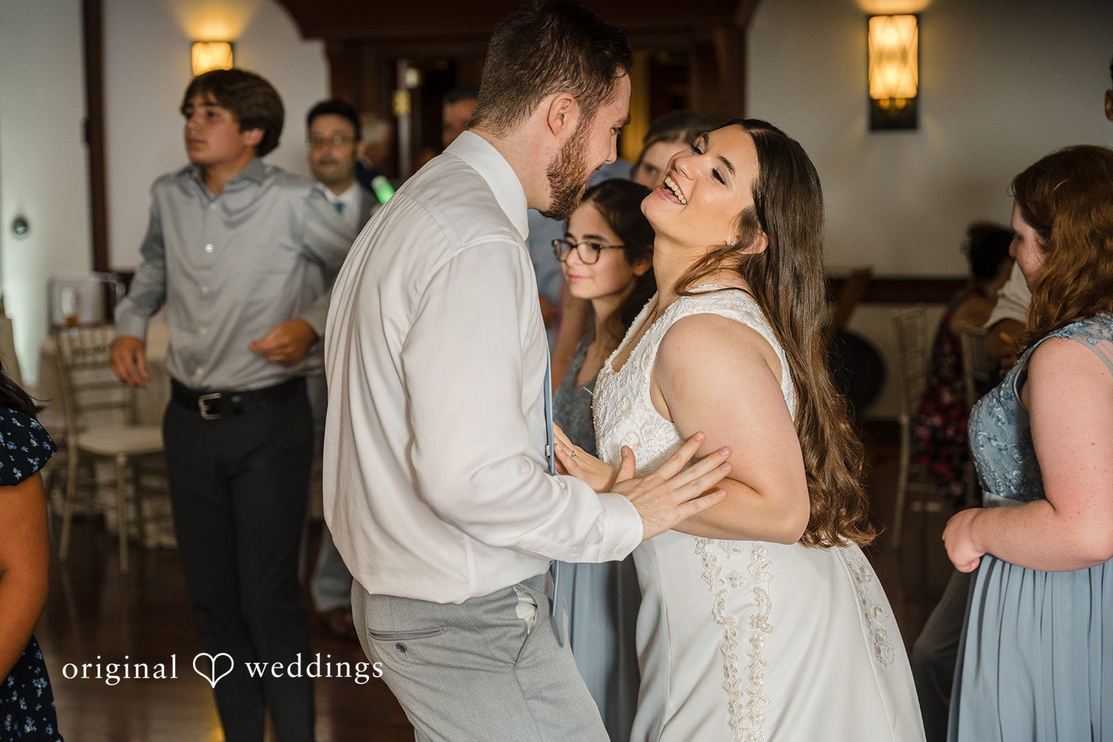 Ava + Zach A smiling bride and groom laughing and dancing together during their wedding reception surrounded by guests.
