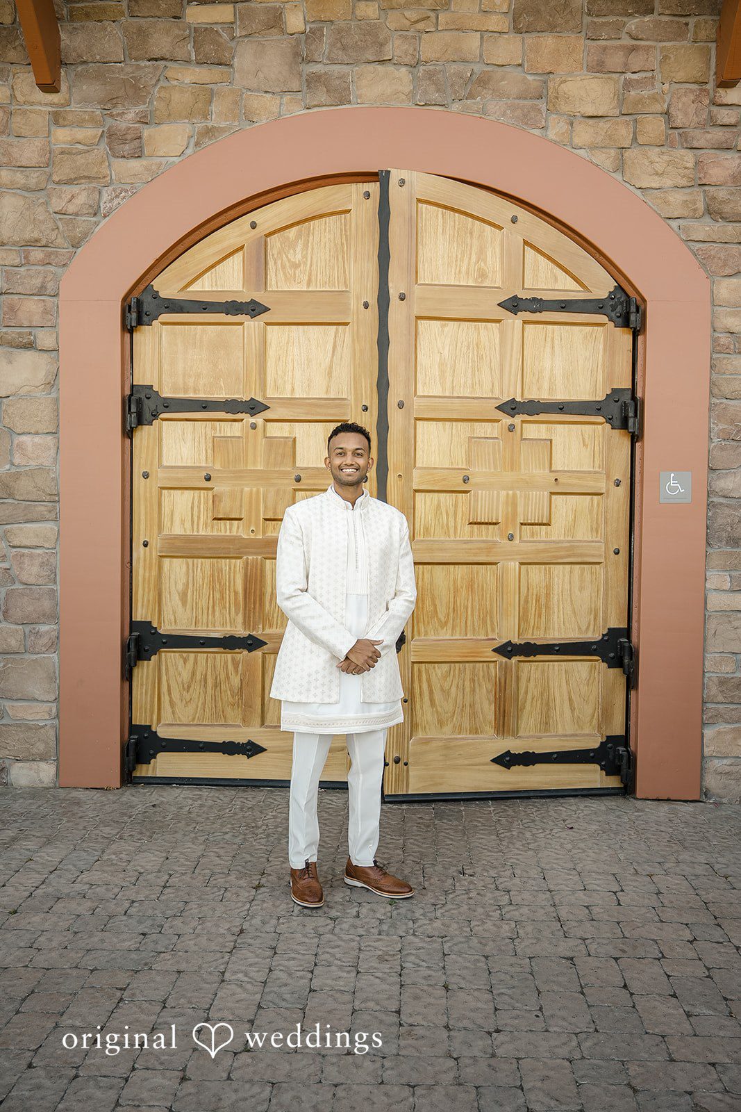 Kathir + Nandita A man dressed in traditional white clothing standing before large wooden doors.