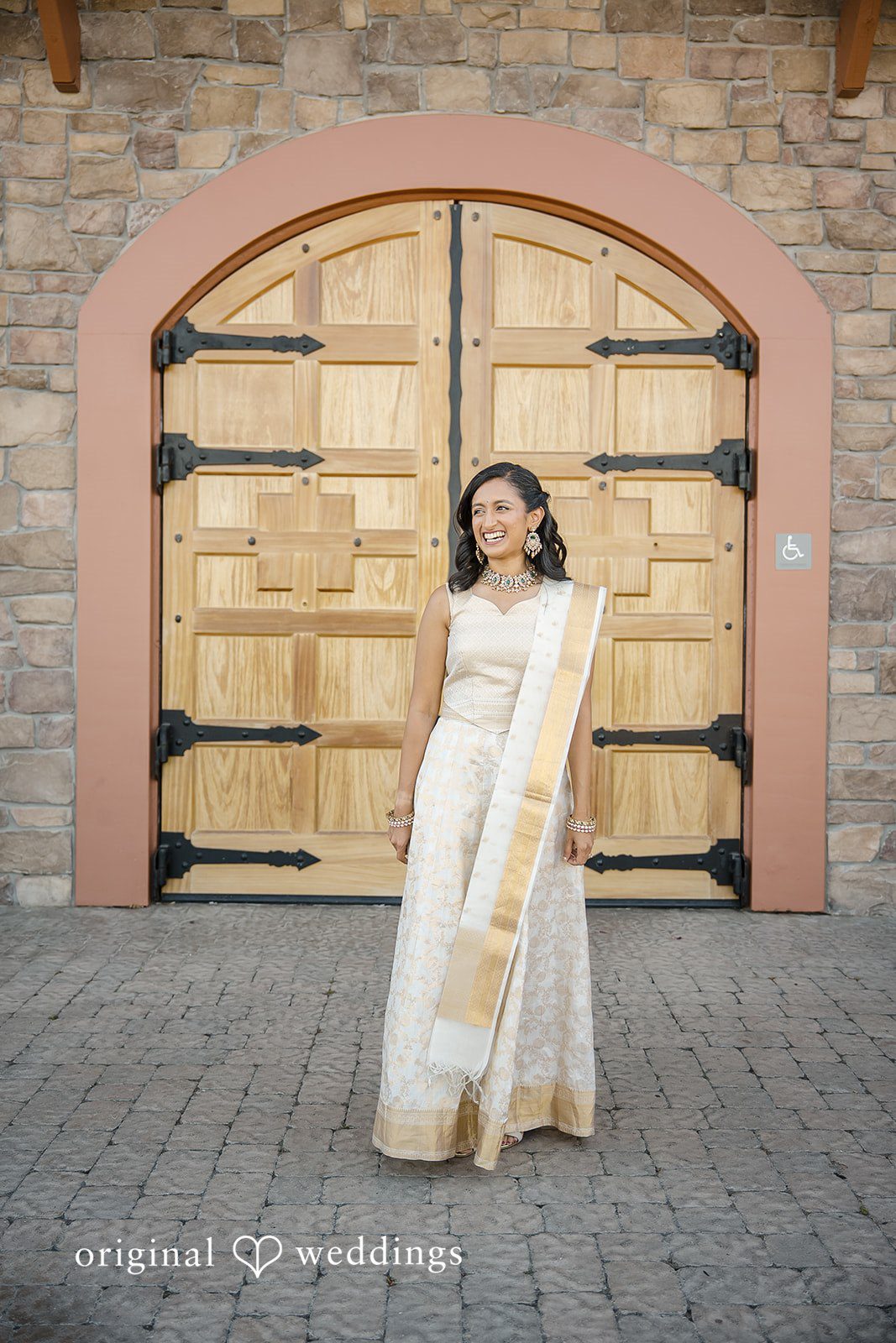 Kathir + Nandita A woman in traditional South Asian attire standing before large wooden doors.